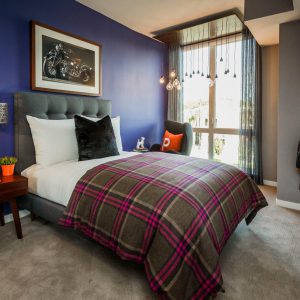 Apartments for Rent in Downtown San Jose, CA - The Pierce - Bedroom with Plush Carpeting, Bed, Nightstand, Wingback Reading Chair, Purple Accent Wall, and Floor-to-Ceiling Windows.