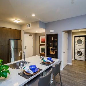 One-Bedroom Apartments in SoFa District, CA - The Pierce - Kitchen with Hardwood-Style Flooring, Kitchen Island, Wood-Tone Flat Panel Cabinets, Stainless-Steel Energy-Efficient Appliances, Solid Surface Stone Countertops, and View of Laundry Closet with Stacked Washer and Dryer.